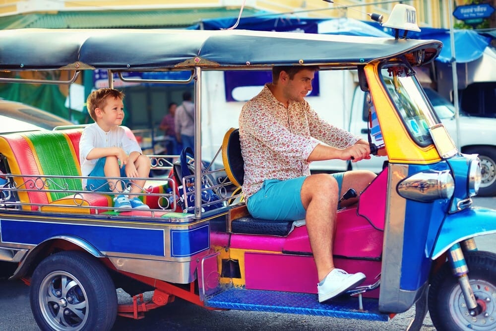 Dad drives colourful tuk tuk with child in back seat in Thailand