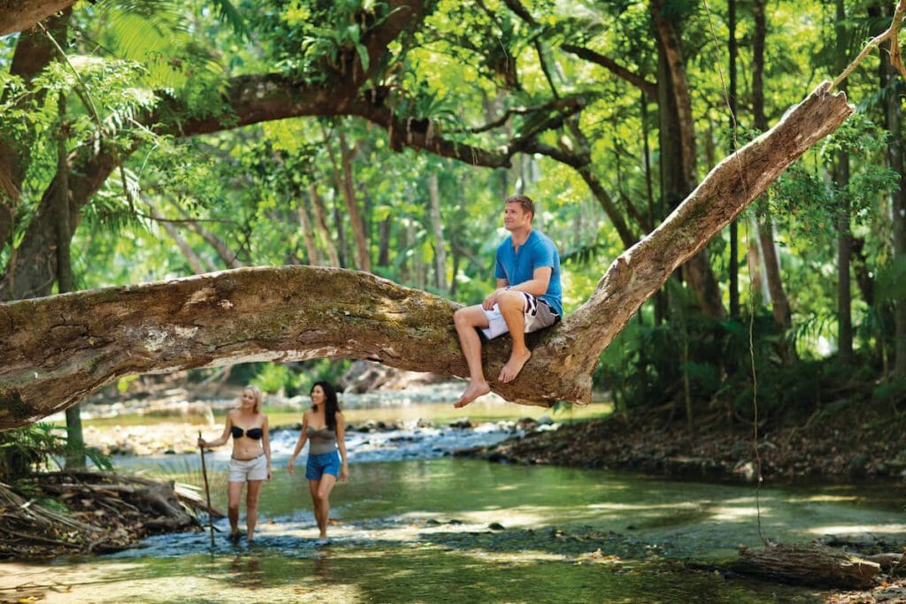 Man sits on tree overhanging creek in the Daintree, two girls in the background