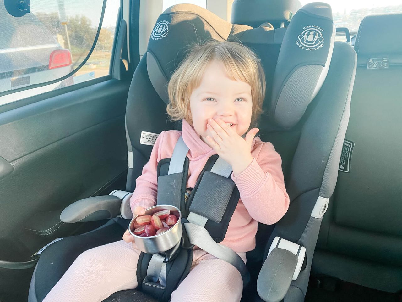 little girl in a pink tracksuit eating grapes while sitting in her car seat in the back of a car