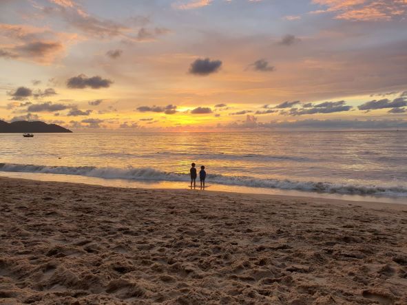 two young kids standing on the waters edge on a beach looking at the sunset