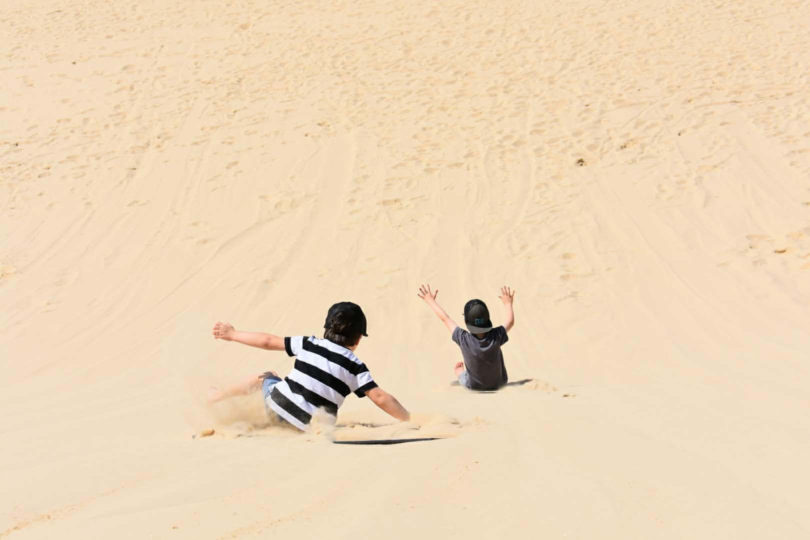 two young boys sliding down a sand dune (image from behind)