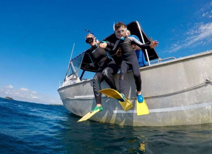 A mother holds her son's hand as they jump from the edge of a tinny into the water in full snorkel gear