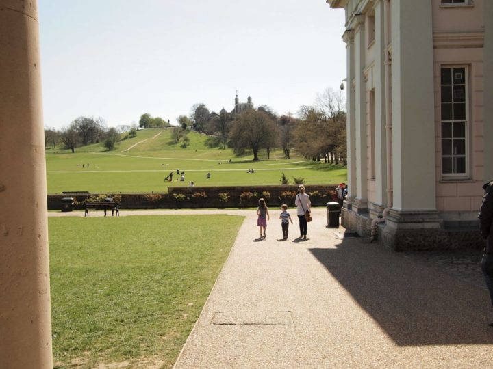 A woman with two young children are walking outside the National Maritime Museum, about to go through Greenwich Park with a view of the Royal Observatory at the top of the hill.