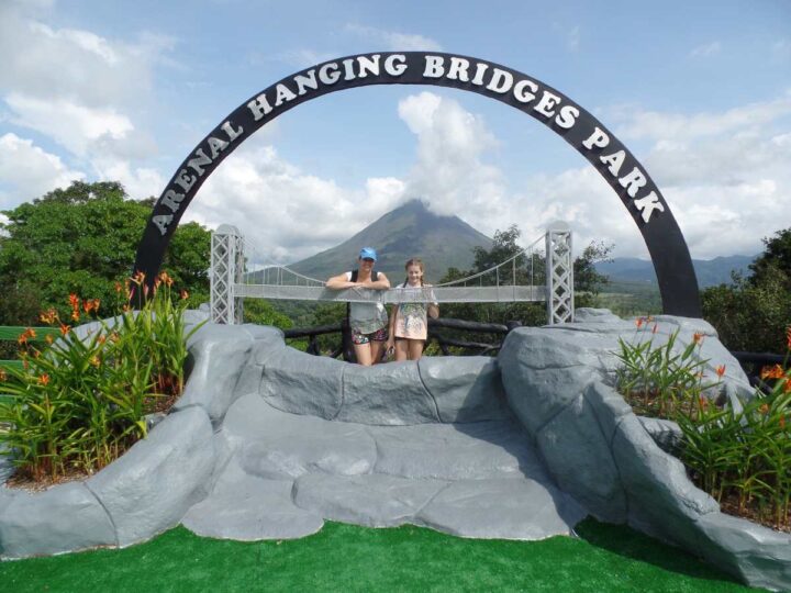 Mother and daughter under Arenal Hanging Bridges Park sign with volcano in the background