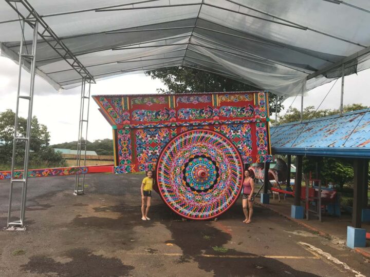 A mother an daughter stand next to the worlds largest ox cart in Costa Rica. The people are only as tall as half the wheel height and the cart is red with lots of colourful flowers painted on it.