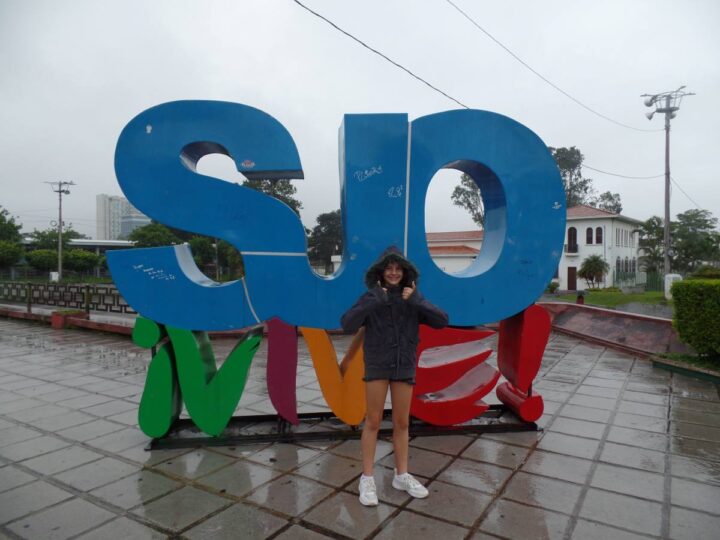 young girl in hooded jacket standing in front of SJO Vive! sign doing a double thumbs up in San Jose, Costa Rica