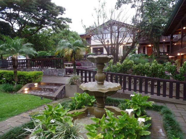 a fountain in the foreground with tropical green gardens in front of a white and dark timber eco lodge in Costa Rica